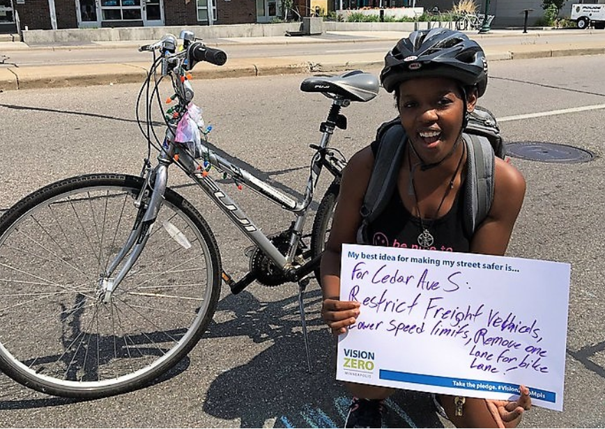 Person holding sign that says "For Cedar Ave S: Restrict freight vehicles, lower speed limits, remove one lane for bike only!" 