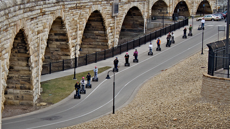 Aerial view of a line of people on Segways with bridge arches in background.