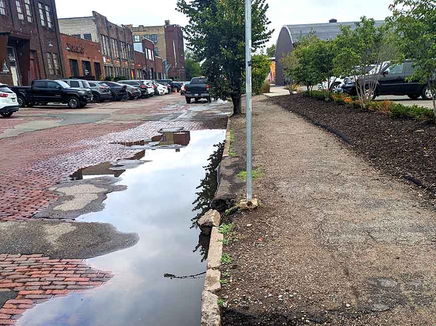 The edge of a brick paved street showing pooled water, a concrete curb broken into many pieces and a deteriorated asphalt sidewalk.