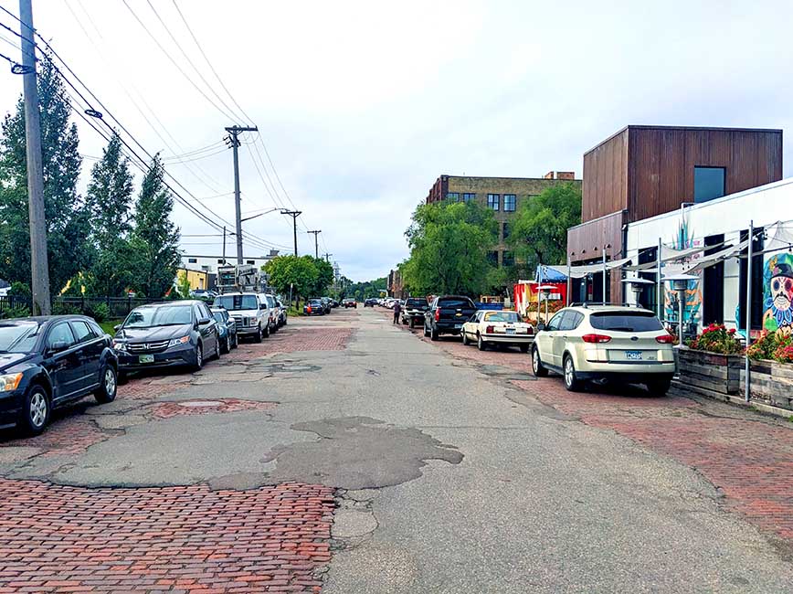 A brick paved street covered with large, overlapping patches of asphalt. Cars are parked along either side of the street.