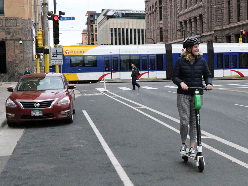 A young woman rides a scooter while the light rail passes by.