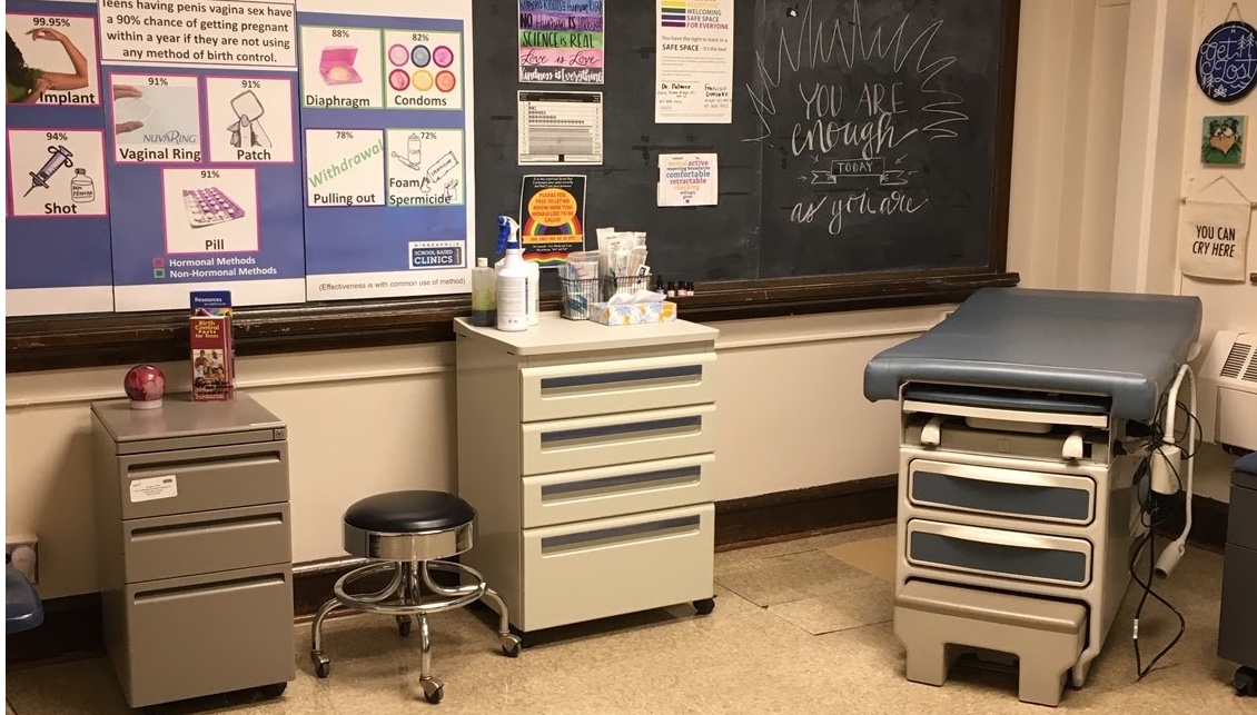 Room with medical exam table, stool and cabinets on wheels, blackboard and posters Room with medical exam table, stool and cabinets on wheels, blackboard and posters
