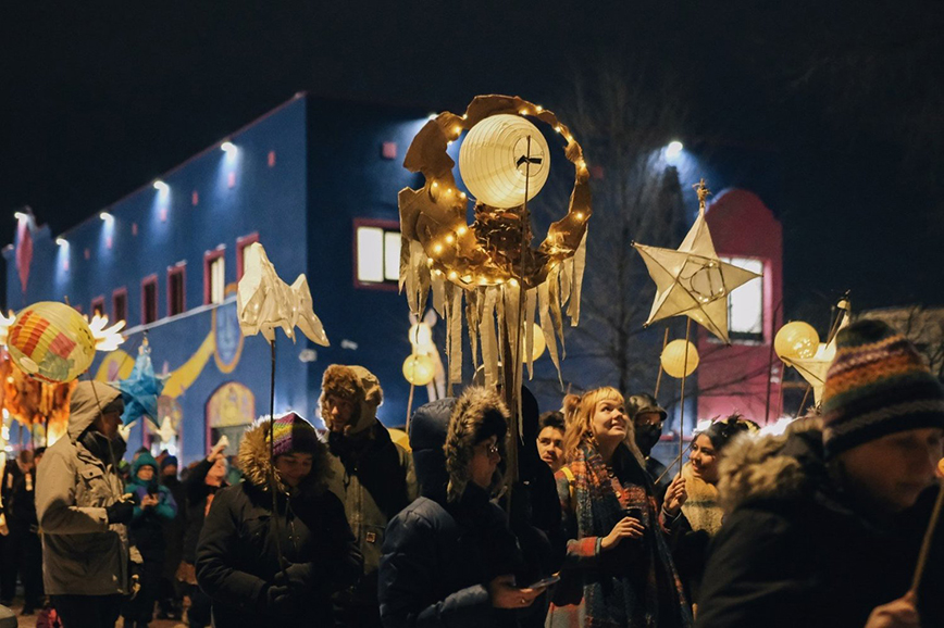 People are parading down Lake Street wearing winter clothing. They carry paper lanterns that illuminate light, in the shapes of a star, moon, and sphe
