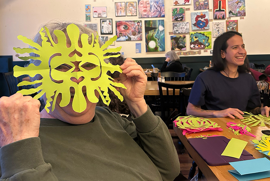 Smiling senior in art-filled cafe holding a yellow hand-cut paper mask in front of their face. Photo credit Witt Siasoco.