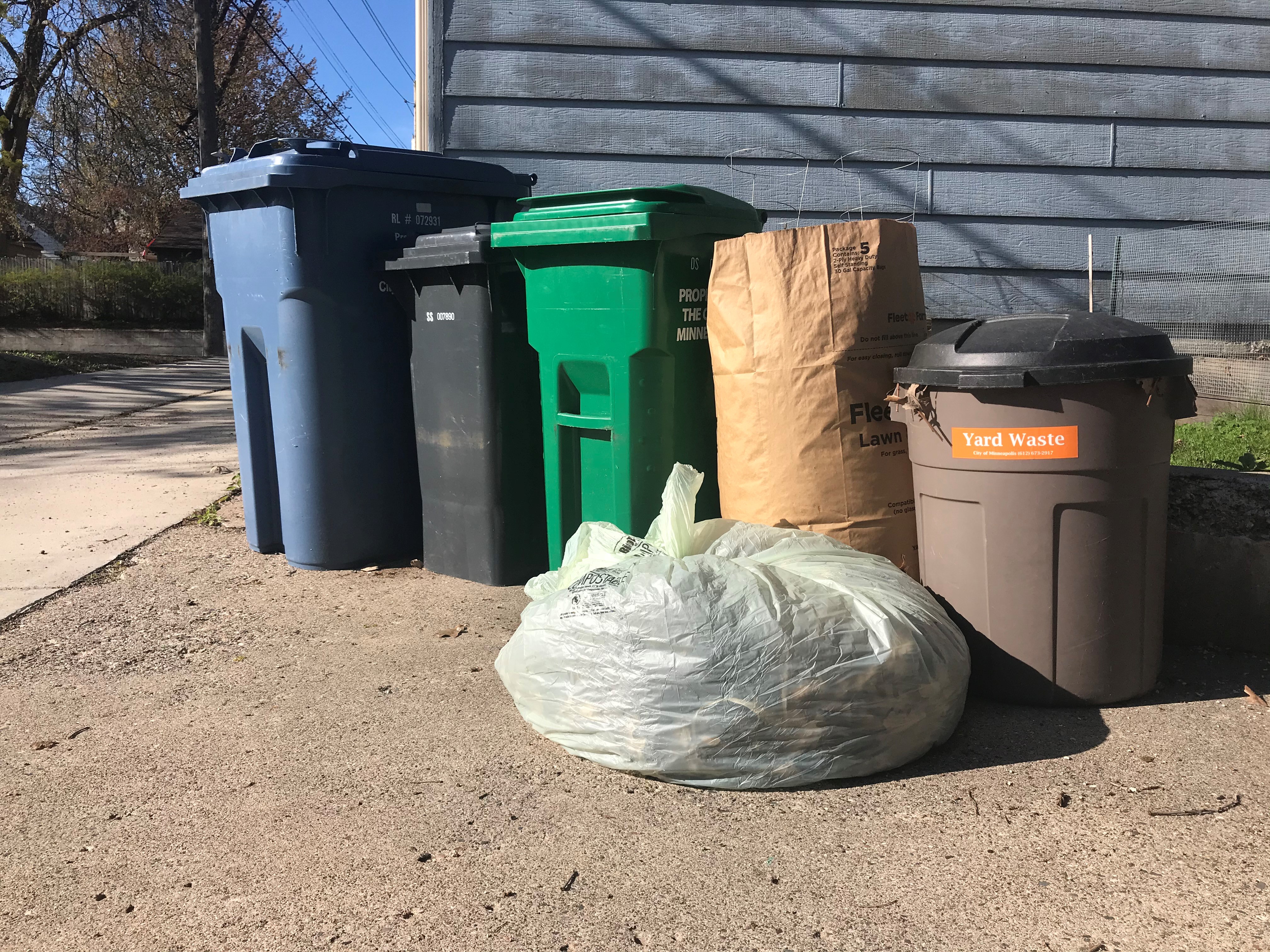 A compostable plastic bag, a compostable paper bag and a reusable yard waste container set out next to the organics, garbage and recycling carts.