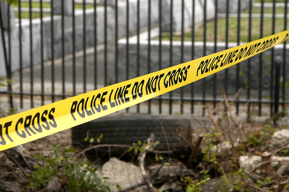 Yellow police tape with black letters saying Police Line Do Not Cross extended across fence.
